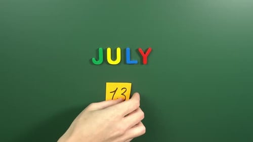 Hand sticking a sticker 13 July calendar day on school board. 13 date of July. Thirteenth day