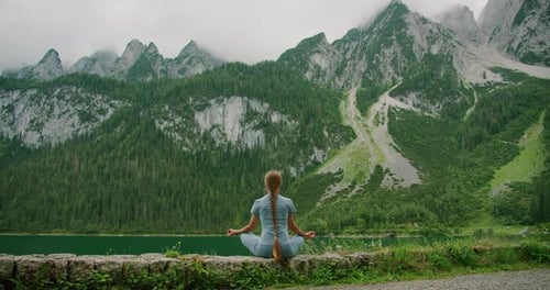 Woman meditates by a serene alpine lake, with dramatic peaks towering in the misty background