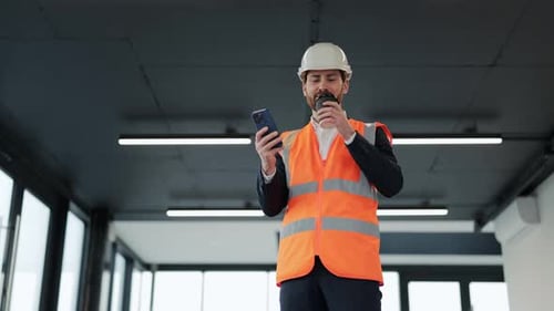 Happy Architect on Coffee Break Browses Smartphone in Building Under Construction