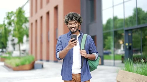 Smiling student with a backpack uses a mobile phone while walking in the campus space near universit