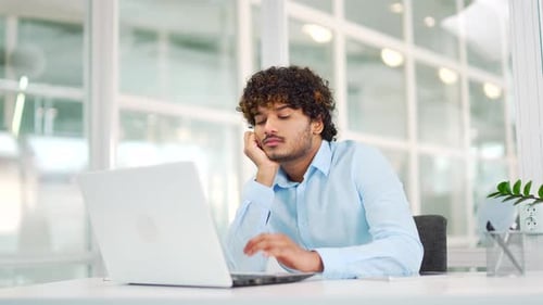 Tired young employee is bored working on laptop while sitting at workplace at desk in modern office.