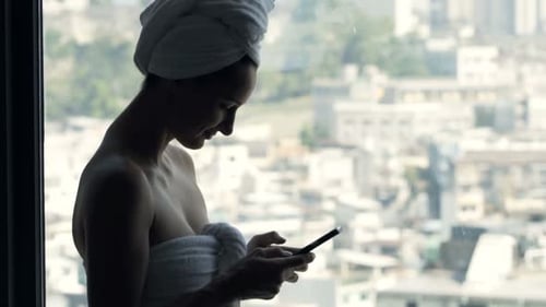 Woman in towel texts on phone by window