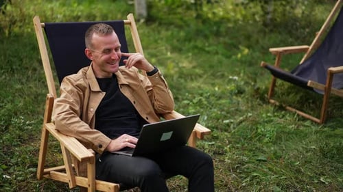 Caucasian man in brown shirt sitting with laptop on his laps in the garden.