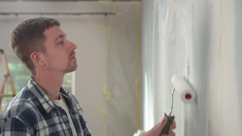 Young Man is Painting Wall with White Paint Using Paint Rollers Against the Backdrop of Window with