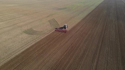 An aerial footage of a tractor working on a large agricultural field, plowing the soil in straight,