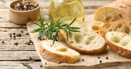 Tranches de ciabatta, huile, grains de poivre et romarin sur une table en bois. La caméra se déplace vers la droite