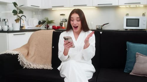 Excited young woman using phone on black sofa