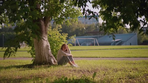 Thoughtful Woman Sitting Under Tree with Stadium Background
