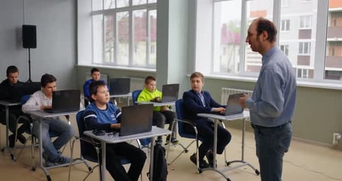 Diversos escolares en el aula, un niño pregunta a los estudiantes que están sentados en mesas con computadoras portátiles