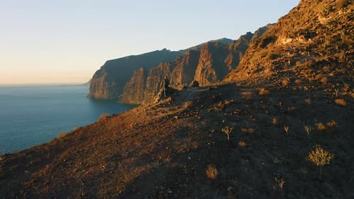 Man with Backpack Climbs the Mountainside and Contemplates Ocean Landscape From Top of Cliff at