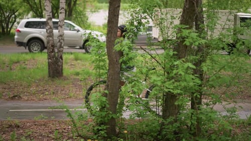 White Woman Pausing With Bicycle Near Road Birch Trees And Passing Cars In Background Contrast