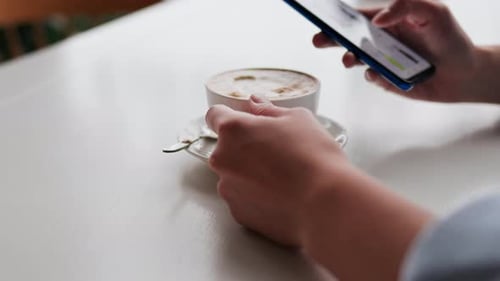 A girl drinks coffee sits in a cafe looks at the smartphone screen choosing clothes in online store