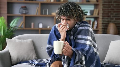 Sick with a cold young adult man coughs while sitting on sofa in living room at home. Upset male