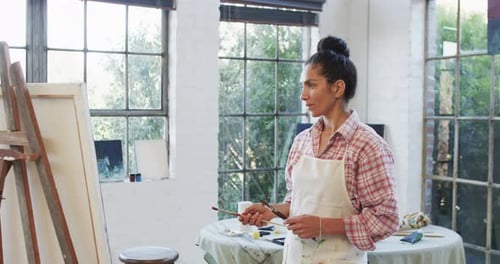 Portrait of biracial female artist in apron holding brushes in studio, slow motion
