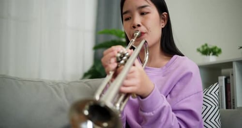 A young woman practices playing the trumpet while sitting on a sofa in a cozy living room.
