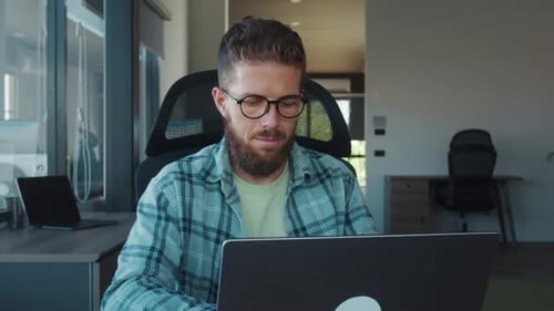 Young Businessman with Eyeglasses and Beard is Sitting at Desk in Modern Office Working on Laptop