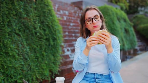Frau beim Mittagessen mit einem Sandwich und Kaffee im Park mit einem Laptop