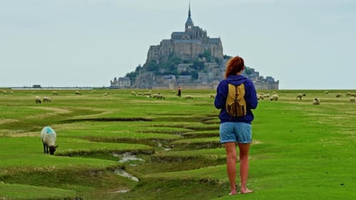A Female with a Backpack Visits the Mont Saint Michel Castle While Walking Through Green Meadows