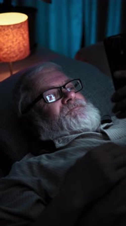 In Dark Room an Elderly Man Puts on His Glasses and Checks the News on His Phone