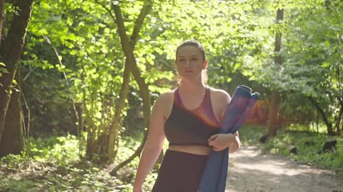 A Woman Holding a Yoga Mat in Her Hands Goes to a Fitness Training in the Park
