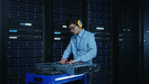 In the Modern Data Center: IT Technician Wearing Protective Headphones Working with Server Racks, o
