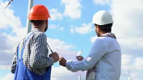 Indian and Caucasian Workers Discussing a Blueprint on a Wind Farm in Front of a Turbine Supplying