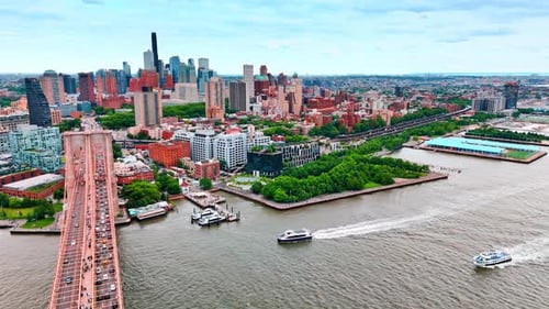 Boats move by the East River towards the Brooklyn Bridge. Plenty of cars go by the bridge.
