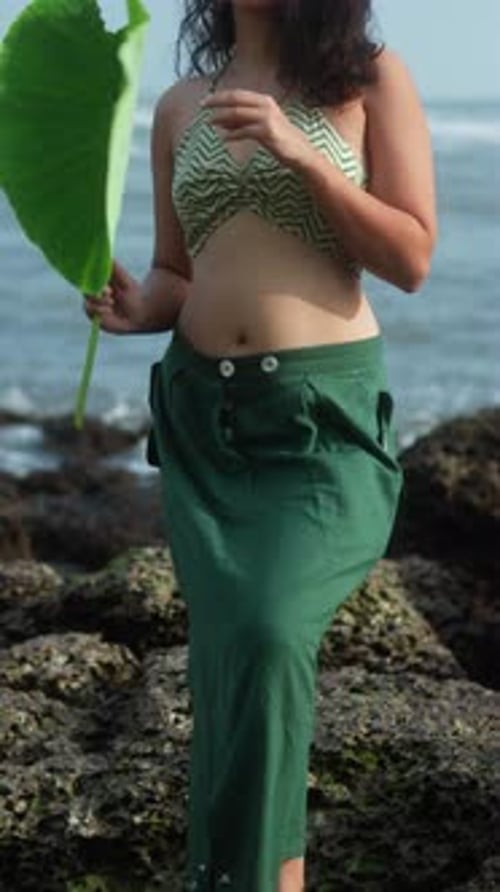 A person holds a green leave at the beach on a sunny day with waves in the background, vertical shot