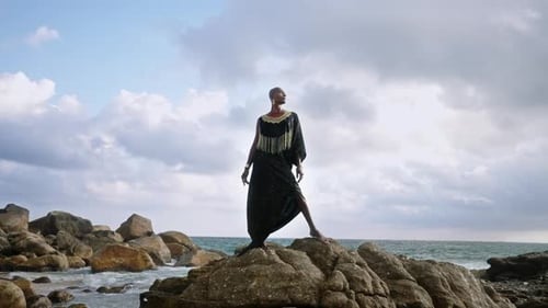 LGBTQ Bipoc Person in Luxury Dress Boutique Golden Jewelry Poses Against Lighthouse on Cliff Ocean
