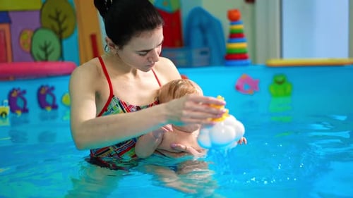 Woman is holding a cute little infant in the swimming pool. Coach is pouring the kid from a toy.