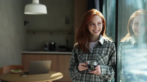 Woman With Red Hair Stands by Window Drinking Coffee