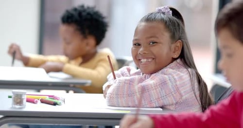In school, smiling girl holding pencil and sitting at desk in classroom