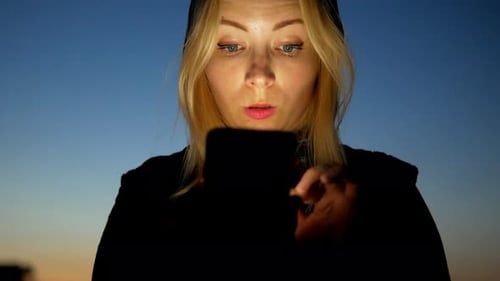 Blond hair girl watches mobile phone with interest at night with sunset sky close-up