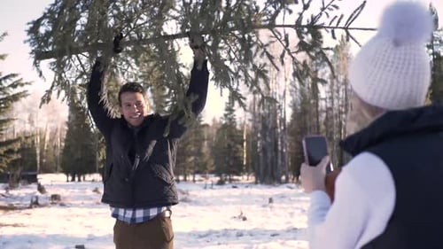 Woman Taking Photo Of Man Lifting Christmas Tree In Forest