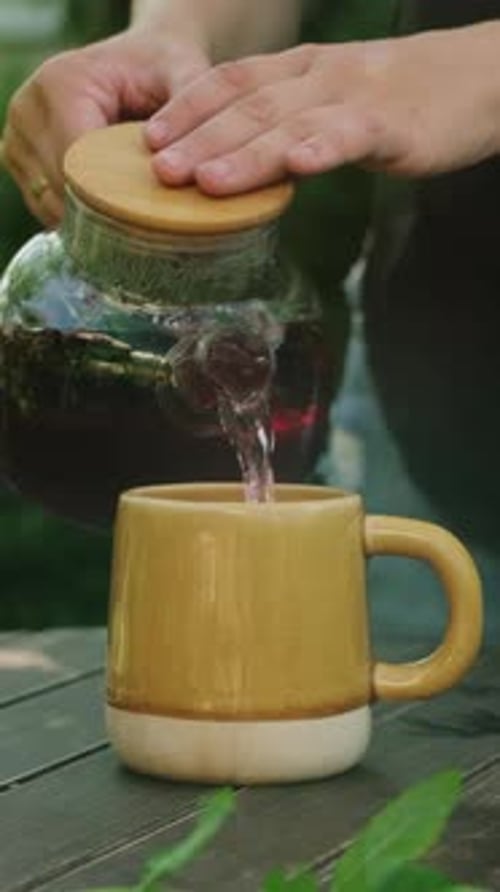 Vertical Video Woman Pouring Red Hibiscus Tea Into Mug on Wooden Table