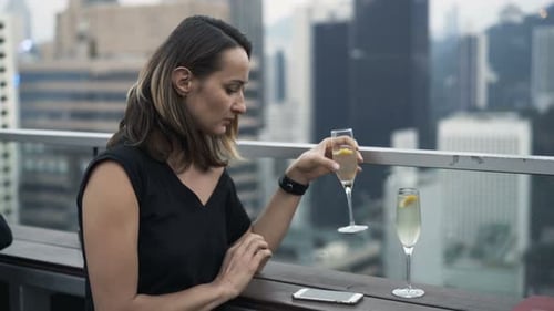 Impatient young woman checks phone and drinks champagne while waiting at rooftop bar