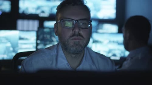 Focused security officer is working on a computer in a dark monitoring room filled with display scr