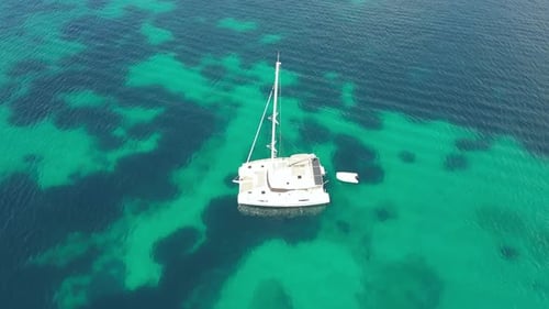 A Drone Flies Around a White Catamaran with a Mast Standing in the Clear Turquoise Sea on a Sunny