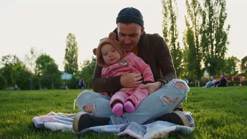 An Adorable Toddler Little Girl in a Pink Suit Sits on a Plaid with Her Dad Spread on the Green