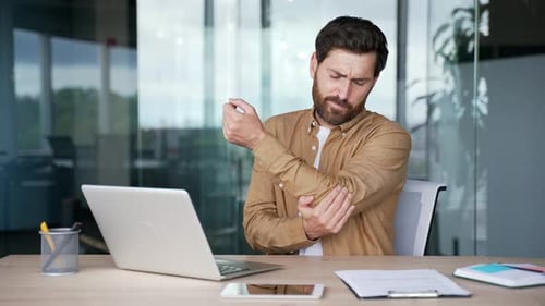 Businessman suffering from elbow pain while sitting at desk at workplace in business office. Upset