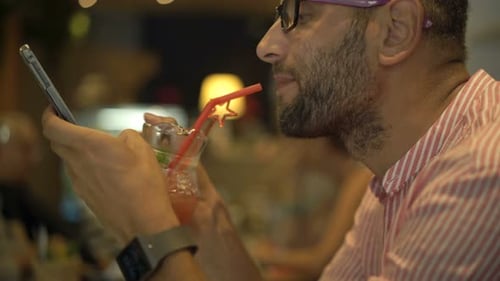 Young Man Texting on Smartphone and Drinking Cocktail in Cafe 30s