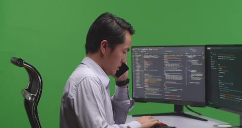 Developer Talking On Mobile Phone While Write Code With Multiple Computer Screens In Green Screen