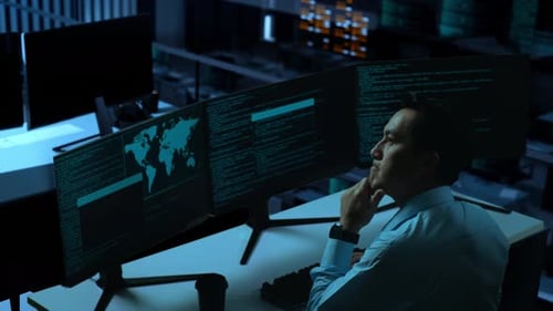 Vista lateral de un hombre asiático pensando mientras escribe código con múltiples pantallas de ordenador en la oficina