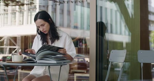 Attractive Young Woman Reading Book While Sitting at Cafe Table Outdoors