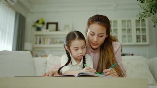 A loving Asian mother helps her young daughter drawing in a coloring book at home.