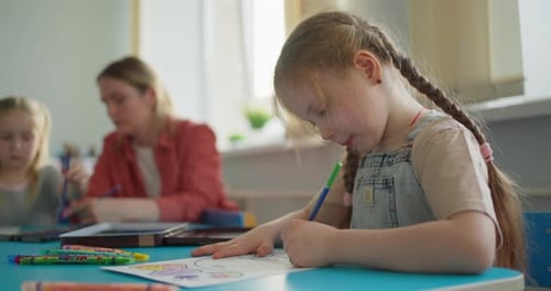 Konzentriertes junges Mädchen mit geflochtenen Haaren beim Färben eines Arbeitsblattes an einem Klassenzimmertisch, während Klassenkameraden und