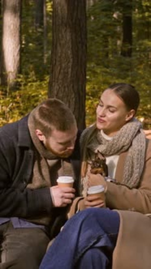 Cariñosa pareja tomando café y besándose en el banco con un cachorro en Fall Park
