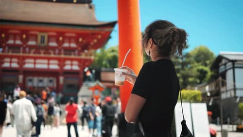 Mujer bebiendo una bebida con una pajita frente a la puerta roja de un templo. Lleva gafas de sol y una o negra