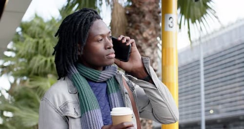 Glancing stop pole, African-American man scrolling phone messages, calling at bus stop with coffee