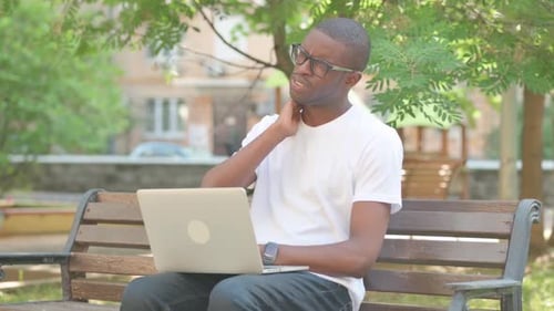 African American Man with Neck Pain Working on Laptop while Sitting on Bench in Park